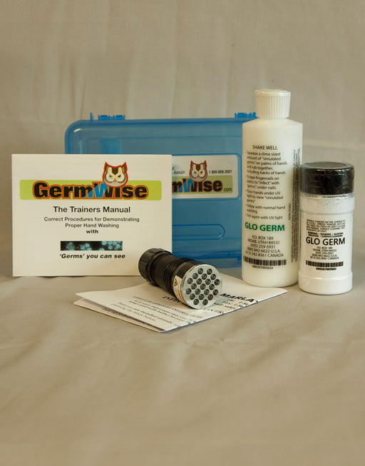 A UV flashlight lies on a beige surface next to a blue plastic case, a manual titled "The Trainers Manual," a bottle, and a jar of "GLO GERM," all related to demonstrating proper handwashing.