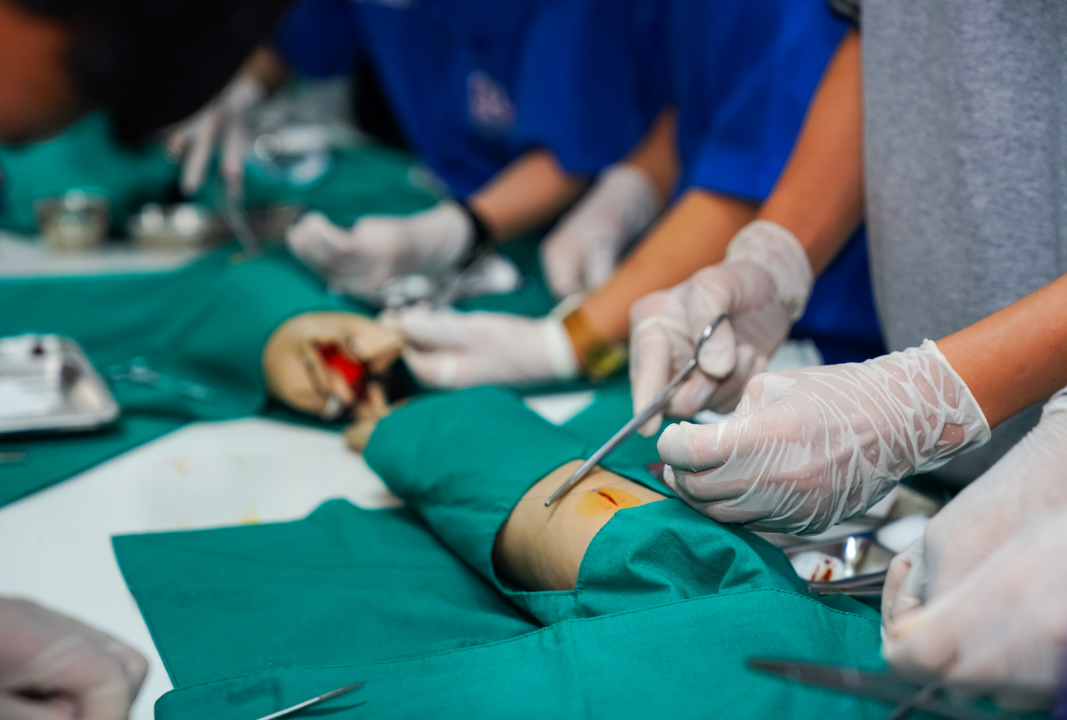 Hands in gloves use surgical scissors on a simulated arm during a medical training exercise.