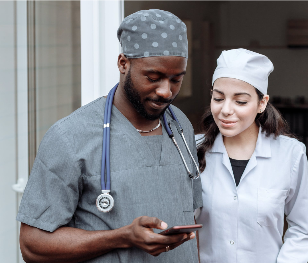 A male and female doctor, wearing scrubs, examine a smartphone; they stand outside a building.