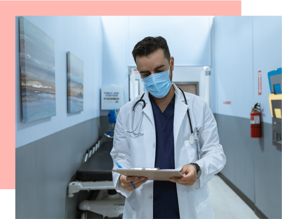 A doctor, wearing a mask, reviews a clipboard in a hospital hallway.
