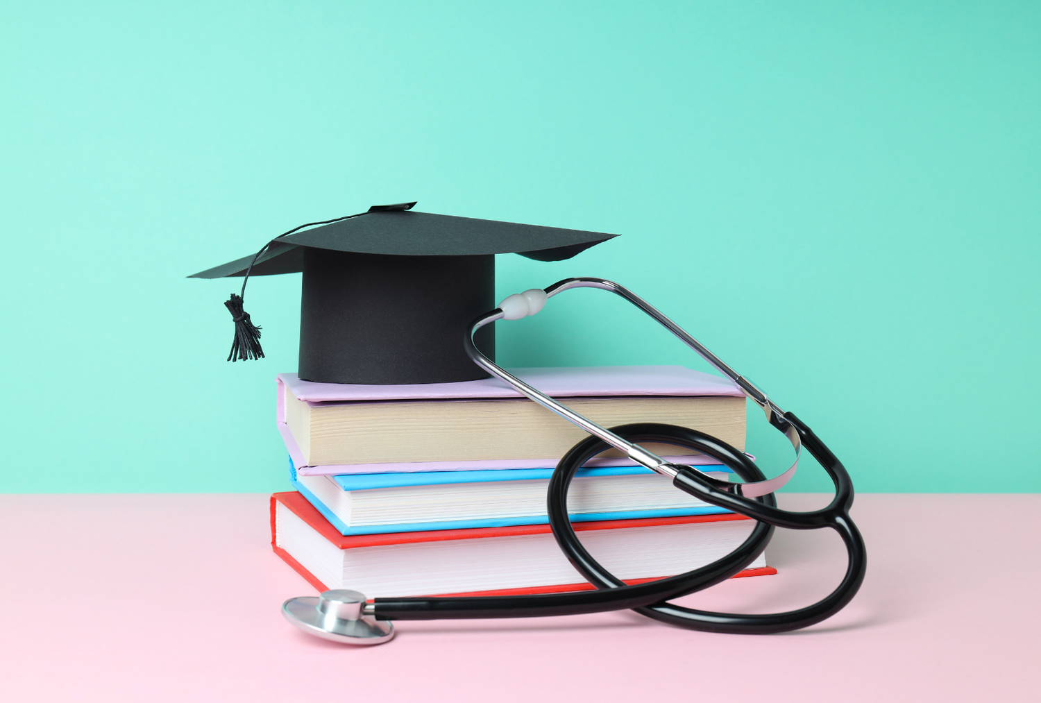A graduation cap rests atop a stack of books, with a stethoscope looped around them. The scene is set against a mint green and pink background.
