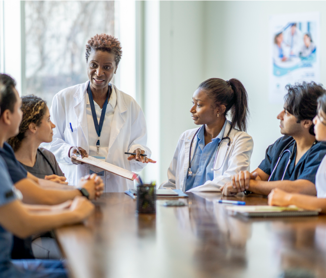 A doctor presents a clipboard during a meeting. Several colleagues, also in medical attire, attentively listen and participate in a conference room setting.