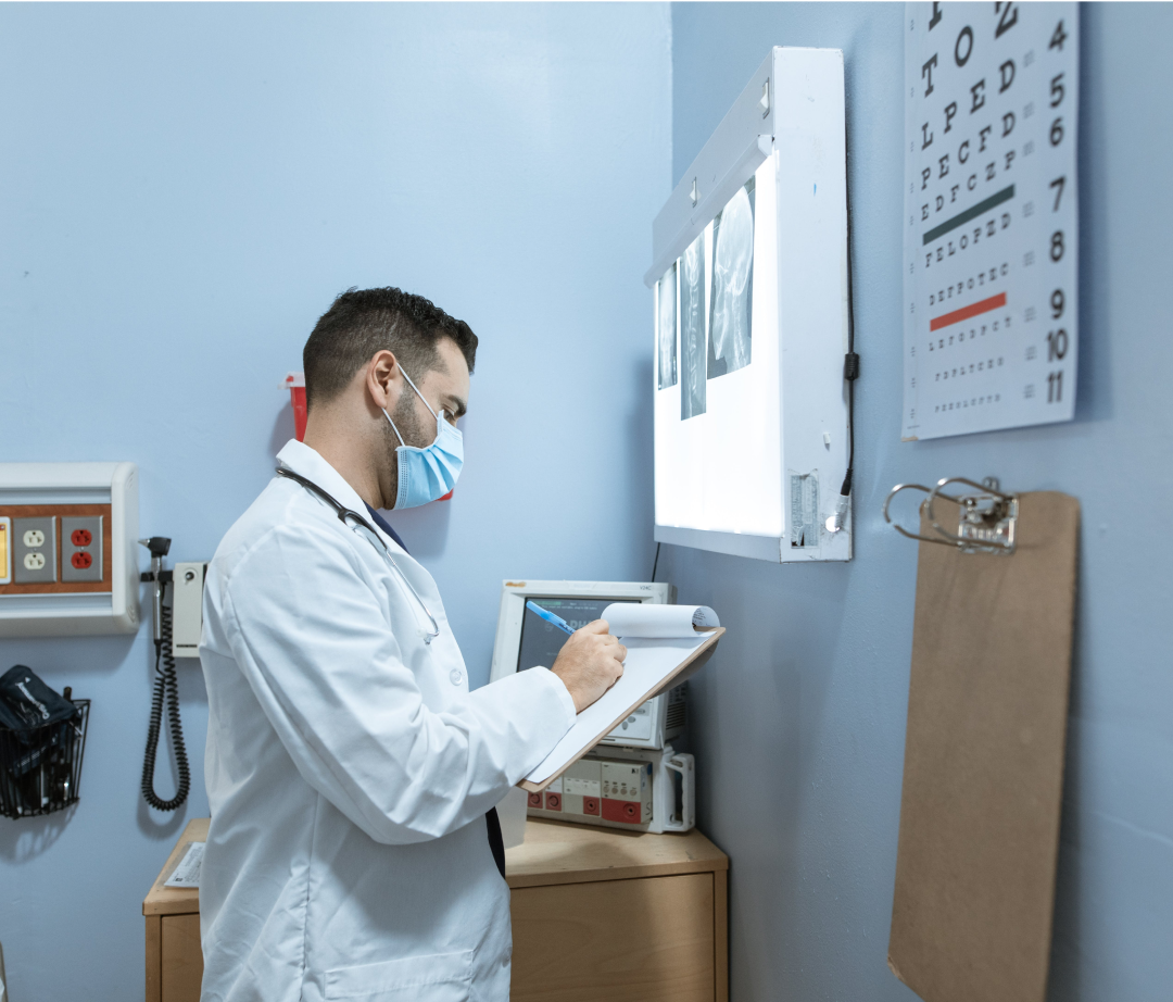 A doctor in a white coat writes on a clipboard. He is wearing a facemask and is reviewing patient x-rays. The setting is a medical examination room.