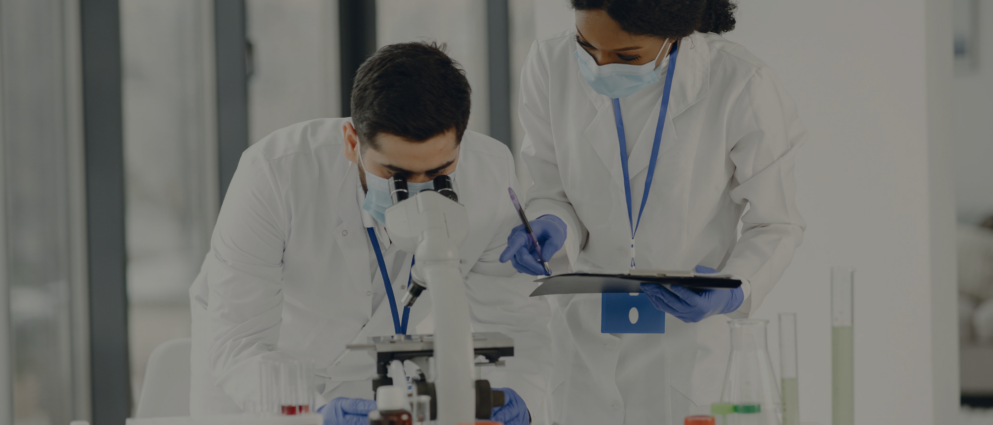 Two scientists, wearing lab coats and masks, collaborate in a laboratory. One uses a microscope; the other reviews a clipboard.