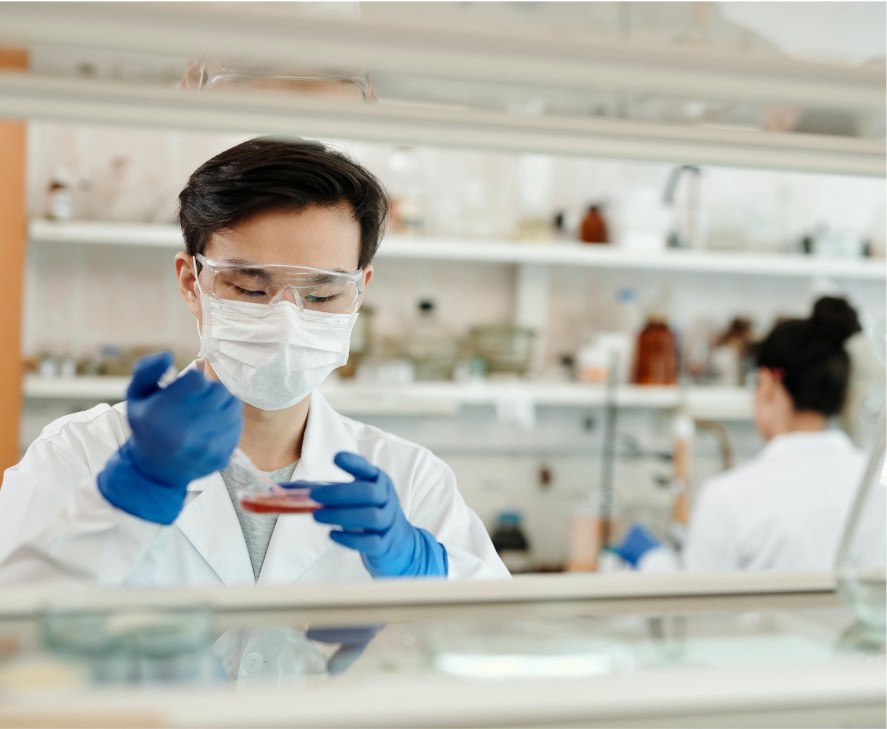 A scientist, wearing gloves and a mask, manipulates a petri dish in a laboratory setting. Another scientist is visible in the background.