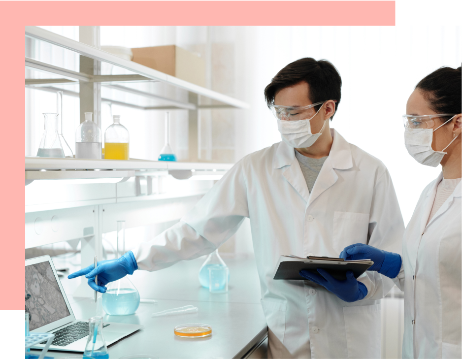 Two scientists, wearing lab coats and protective gear, examine a laptop displaying a microscopic image in a laboratory setting.