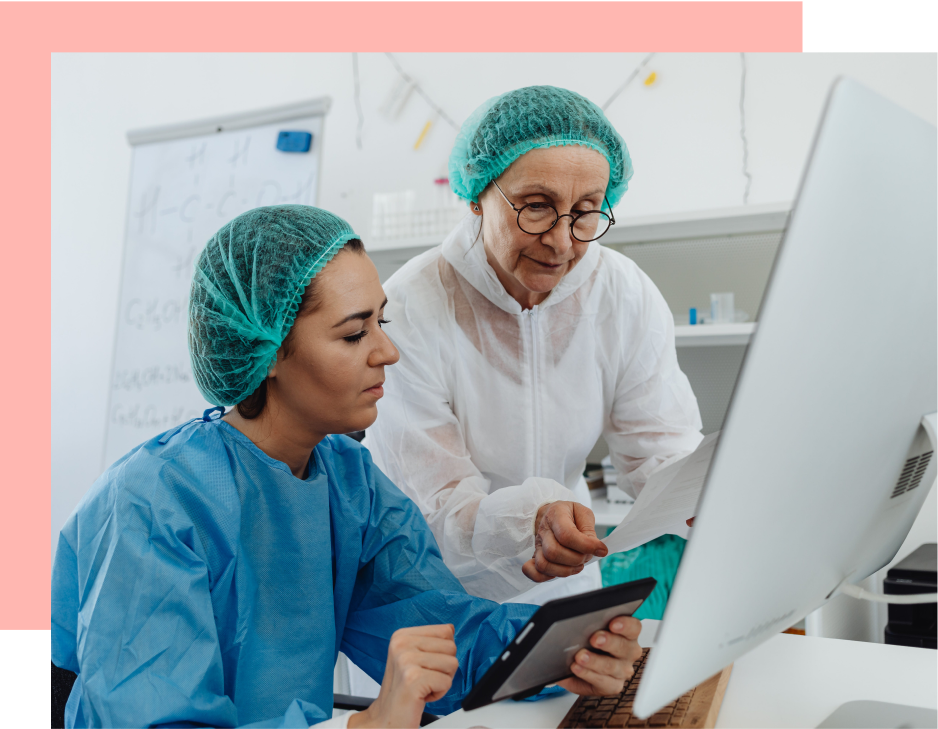 Two lab technicians, wearing protective gear, review documents and a tablet at a computer in a laboratory setting.