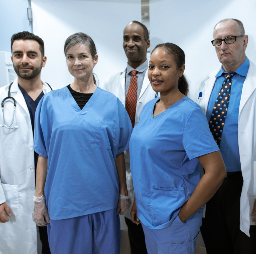 A diverse group of five medical professionals stand together in a clinical setting. They are wearing scrubs and lab coats, and appear confident and composed.