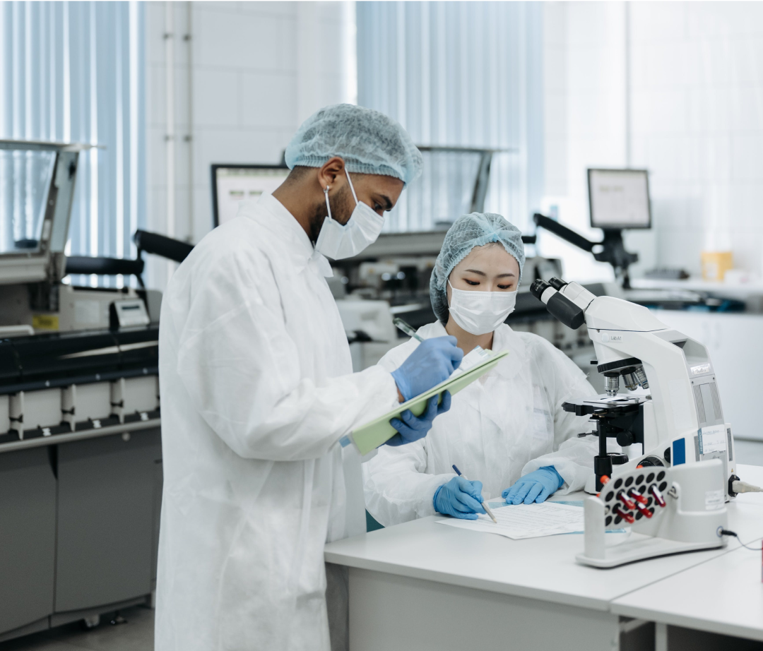 Two scientists, wearing lab coats and masks, review documents near a microscope in a laboratory.