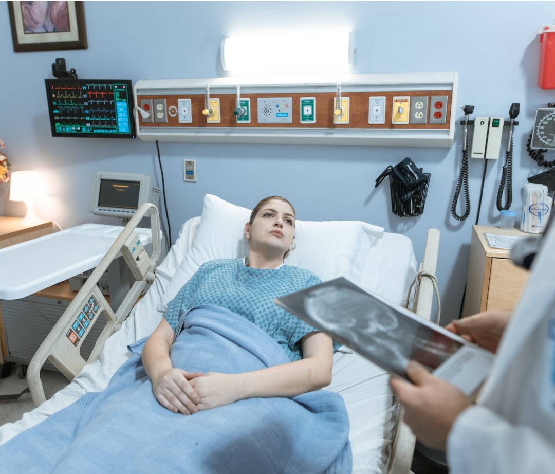 A young woman lies in a hospital bed, passively receiving medical attention. A doctor holds up what appears to be a medical scan. The setting is a hospital room.