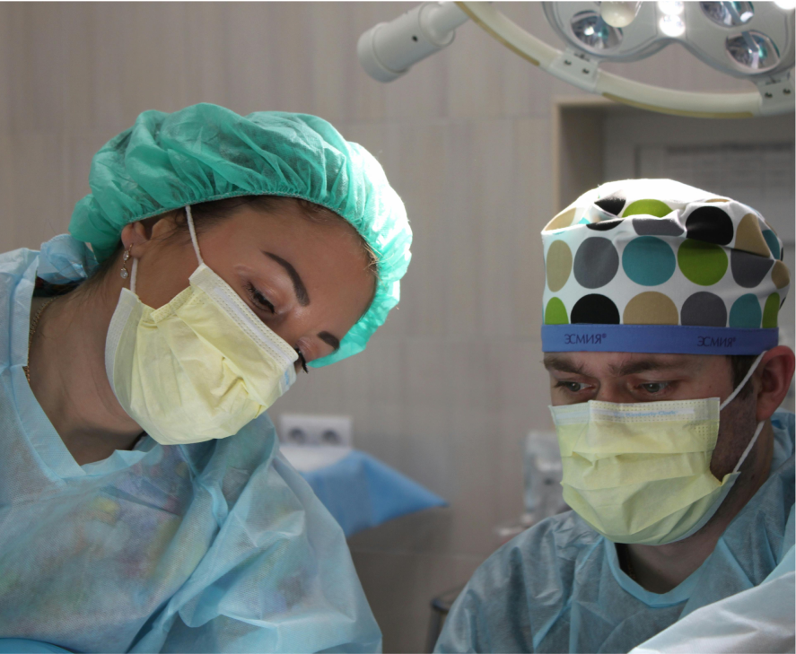 Two masked surgeons, wearing surgical caps, are leaning in; one's cap is teal, the other's is multicolored with the word "ЗСМІЯ" printed on a blue band.