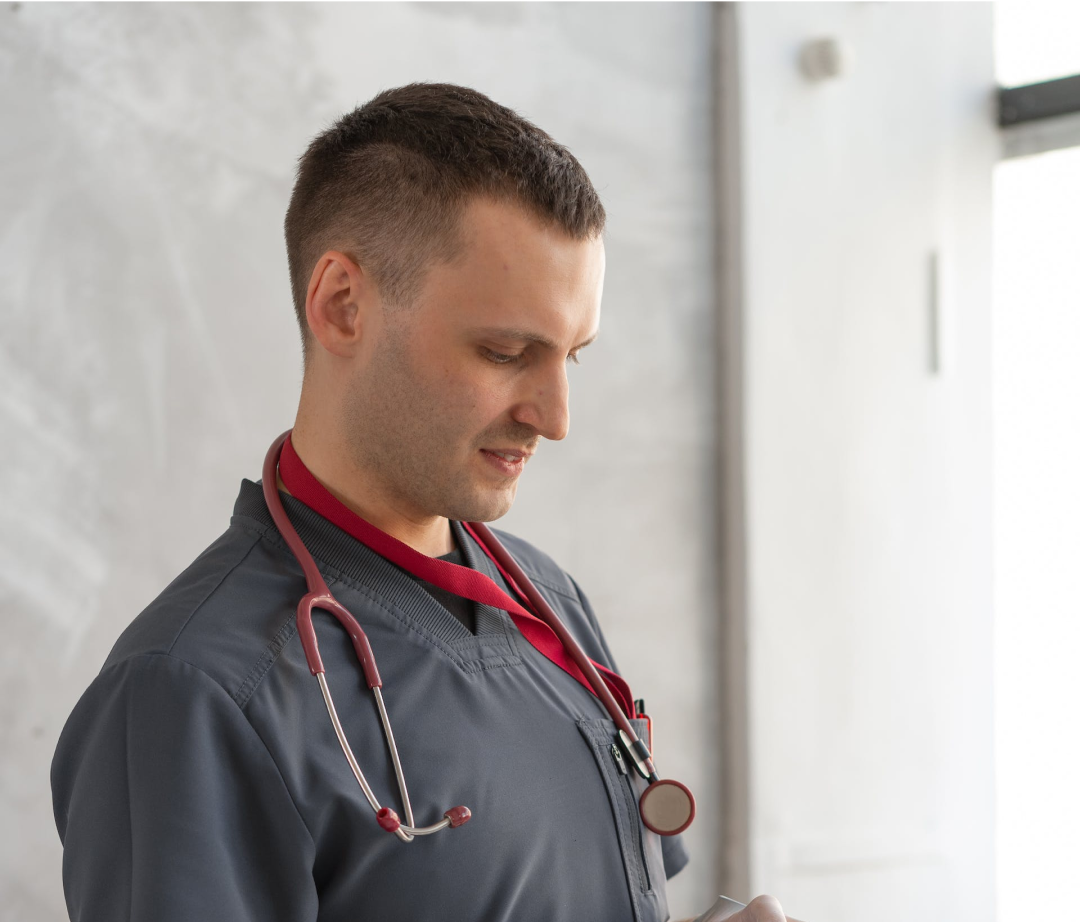A male doctor, wearing a grey scrub and a burgundy stethoscope, looks down in a bright, possibly clinical, setting.