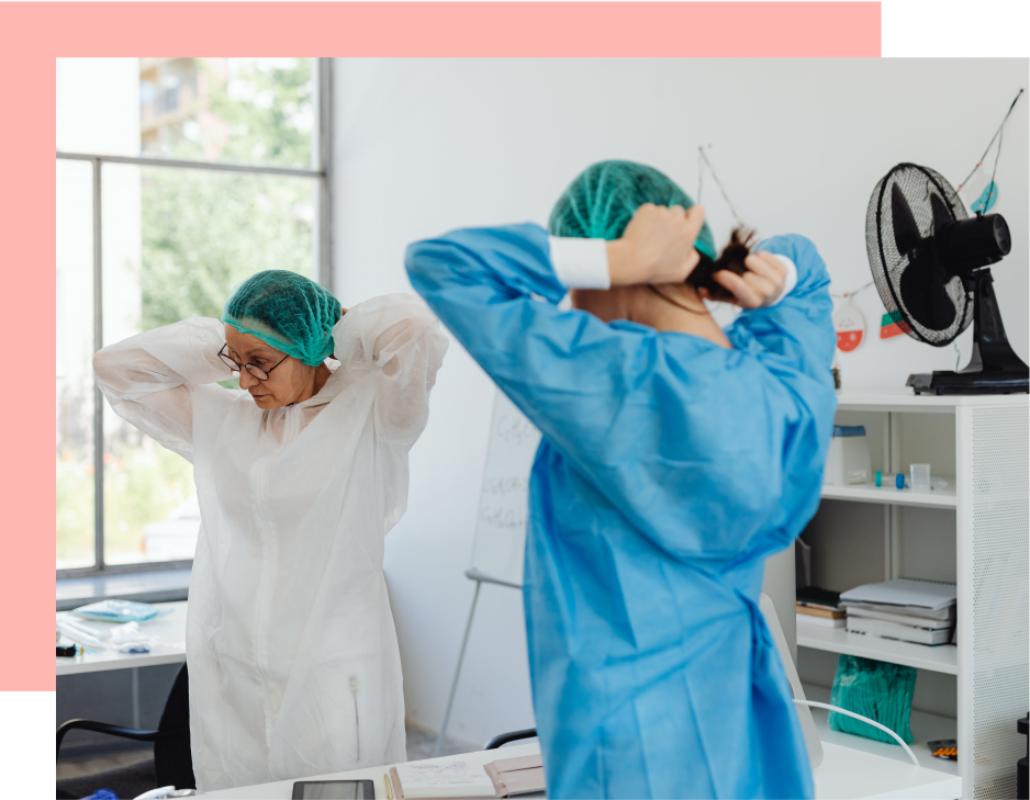 Two people don protective gowns and caps in a bright room. A fan is visible in the background.
