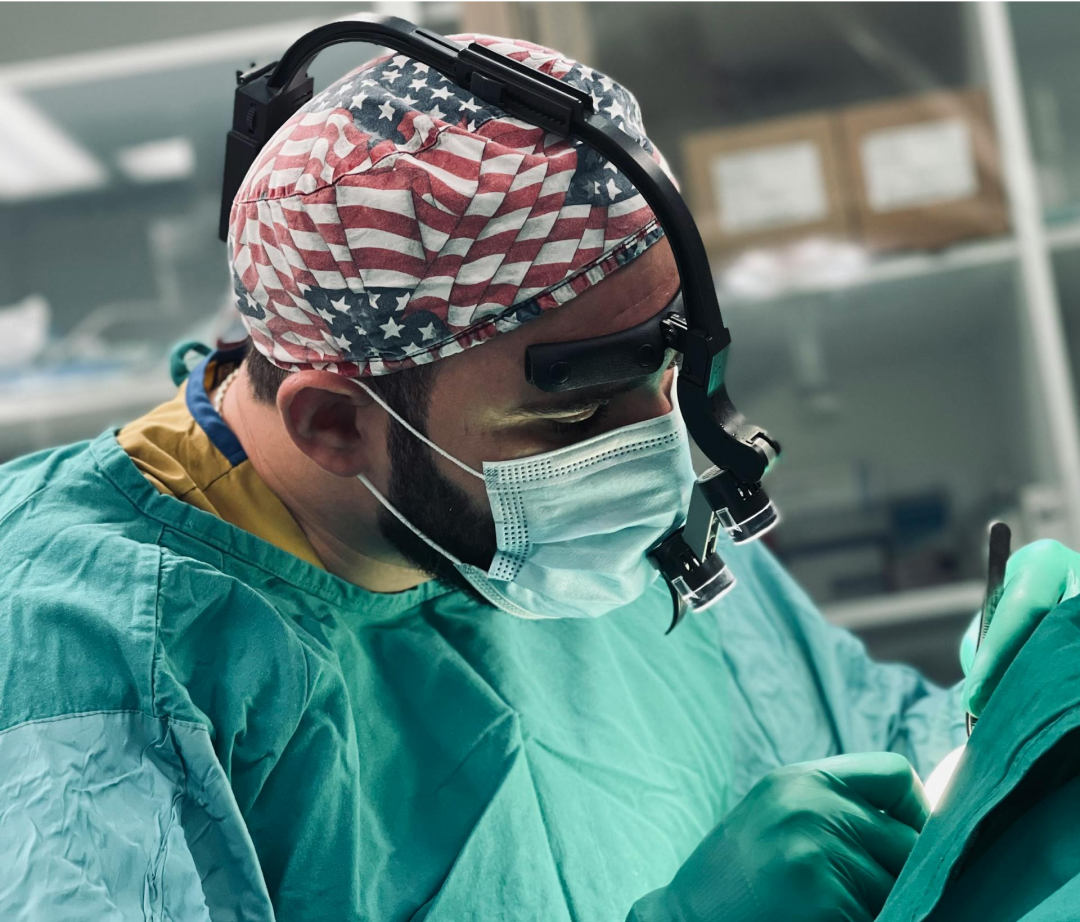 A surgeon, wearing a USA-flag patterned surgical cap and loupes, performs a procedure in an operating room.