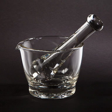 A clear glass mortar and pestle rest on a dark surface; the pestle sits inside the bowl of the mortar.
