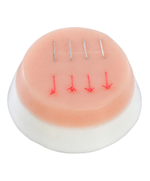 A pale pink, dome-shaped training pad displays four simulated surgical sutures; the sutures are represented by small, thin metal pins and red markings on the surface.