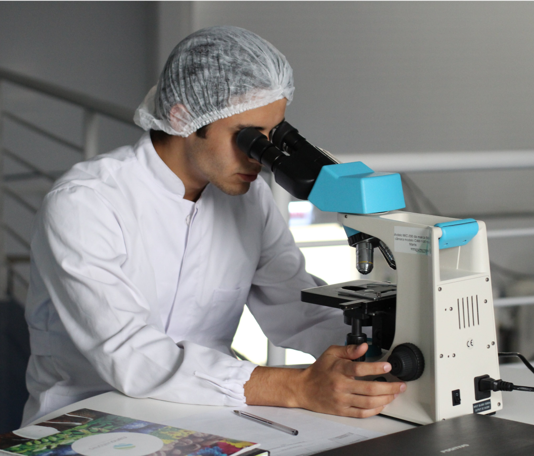 A scientist, wearing a lab coat and hairnet, uses a microscope in a laboratory setting. Modelo MC-1005 da marca Euromex, Câmara modelo MC-1005, Marca Euromex.
