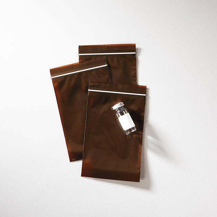 Three brown resealable bags lie on a white surface; a small vial with a blank label rests on top.