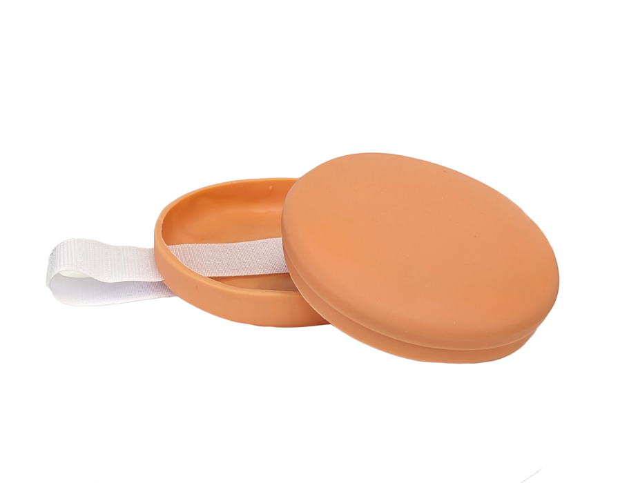 A peach-colored, round sponge sits in a similarly colored, slightly smaller, lidded container; a small white ribbon is attached to the container. The scene is set against a plain white background.