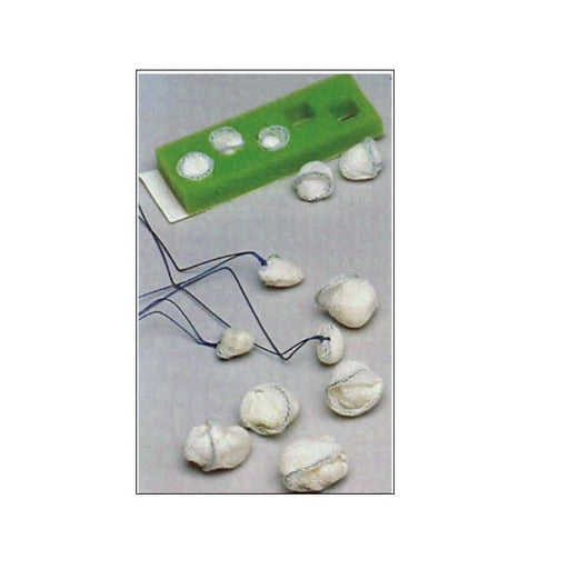 Small, gauze-like objects with attached strings are scattered on a surface near a green rectangular mold containing similar objects.