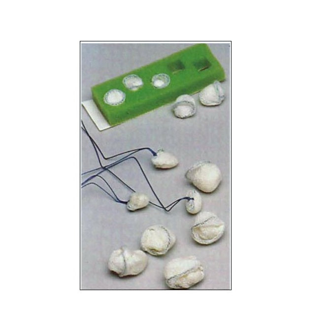 Small, gauze-like objects with attached strings are scattered on a surface near a green rectangular mold containing similar objects.