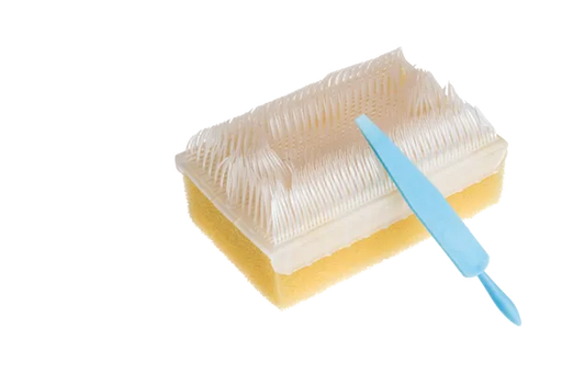 A rectangular sponge brush, featuring a dense, white bristle top, rests on a surface. A light blue cleaning tool lies atop the brush.