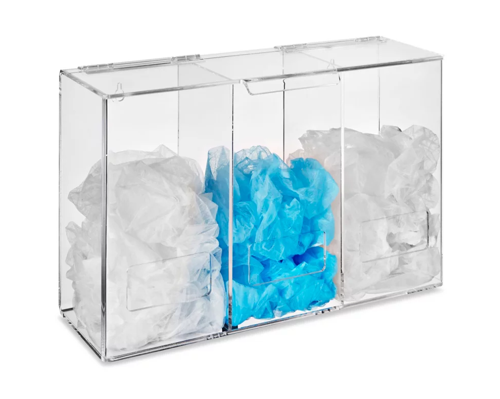 A clear acrylic box holds piles of white and light blue disposable coverings; it's likely a dispenser in a clinical or sanitary setting.