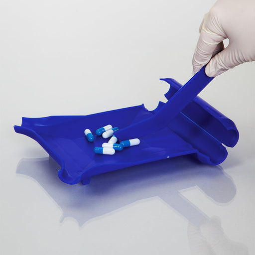 A blue plastic pill tray holds several blue-and-white capsules. A gloved hand uses a blue spatula to move the pills. The setting is a clean, light-colored surface.