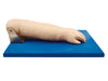 A realistic arm model rests on a blue base. The arm is positioned as if lying on a flat surface. The model is likely used for medical training.