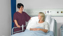 A nurse assists a patient resting in a hospital bed. The nurse is in maroon scrubs; the patient is in a light-colored gown. The setting is a hospital room.