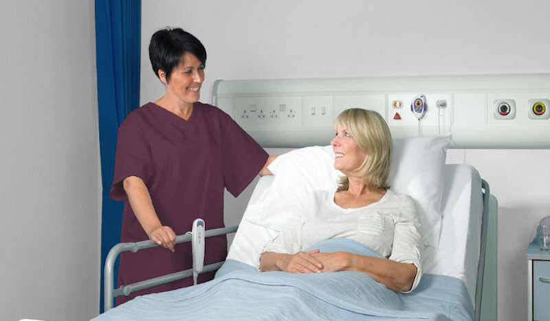 A nurse assists a patient resting in a hospital bed. The nurse is in maroon scrubs; the patient is in a light-colored gown. The setting is a hospital room.