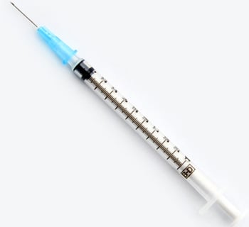A disposable syringe rests on a white surface. It is clear plastic with a dark-colored plunger and a light-blue plastic hub. Measurements are printed along the barrel. The needle is attached.