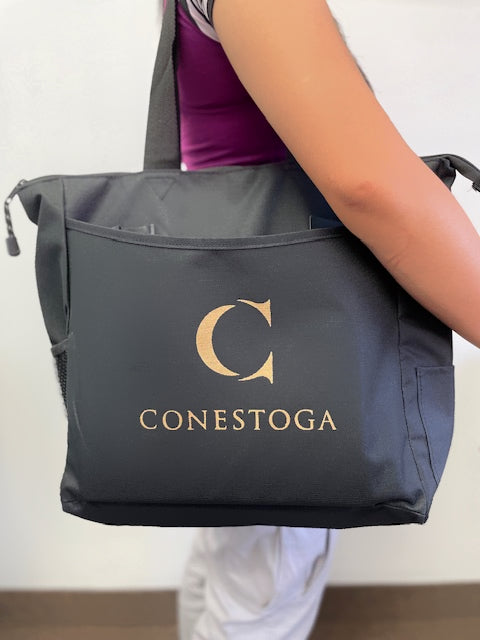 A black tote bag, featuring a gold "C" and "CONESTOGA," is being carried by a person. The bag has side pockets and is worn in an indoor setting.