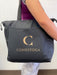 A black tote bag, featuring a gold "C" and "CONESTOGA," is being carried by a person. The bag has side pockets and is worn in an indoor setting.