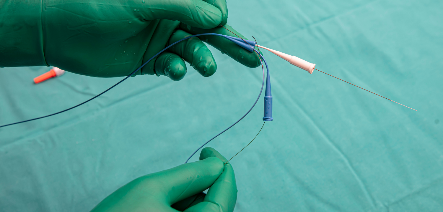 Gloved hands manipulate a thin, wired medical device with blue and white components on a teal surface.