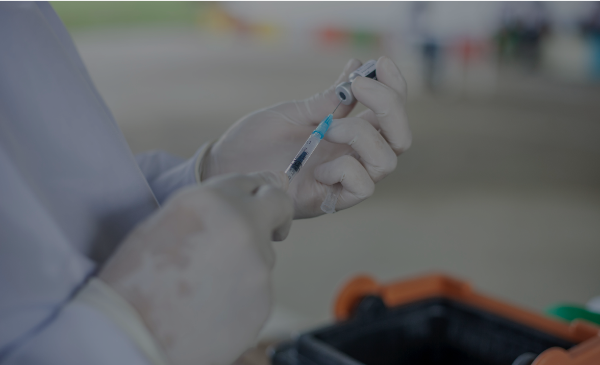 Gloved hands draw vaccine from a vial into a syringe. This occurs indoors in a blurry, possibly medical, setting.
