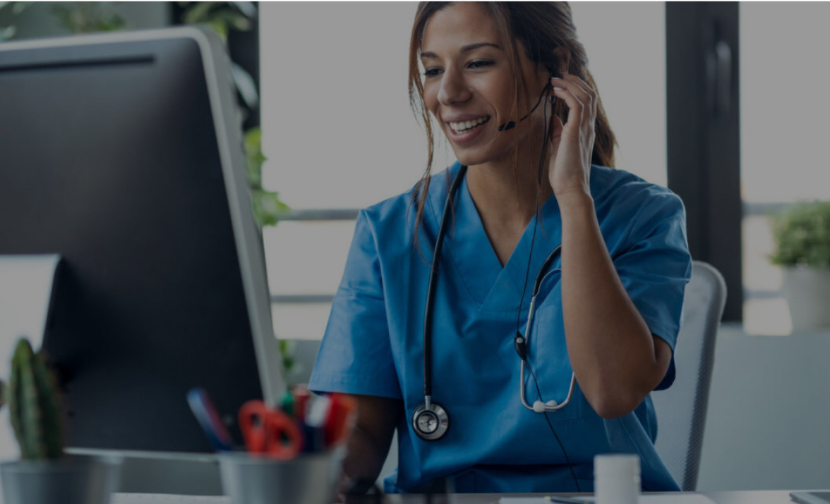 A nurse uses a headset while working at a computer in a brightly lit office.