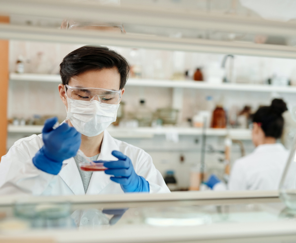 A scientist, wearing gloves and a face mask, manipulates a petri dish in a laboratory setting. Another scientist is visible in the background.