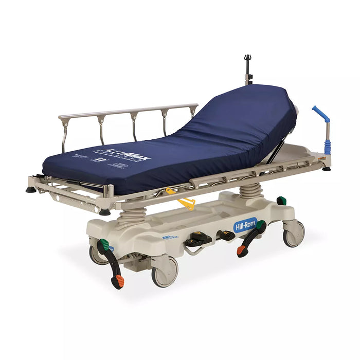 A stretcher rests on wheels, featuring a dark blue mattress; it's in a sterile environment. The text on the mattress reads "AirMax." The stretcher is labeled "Hill-Rom."