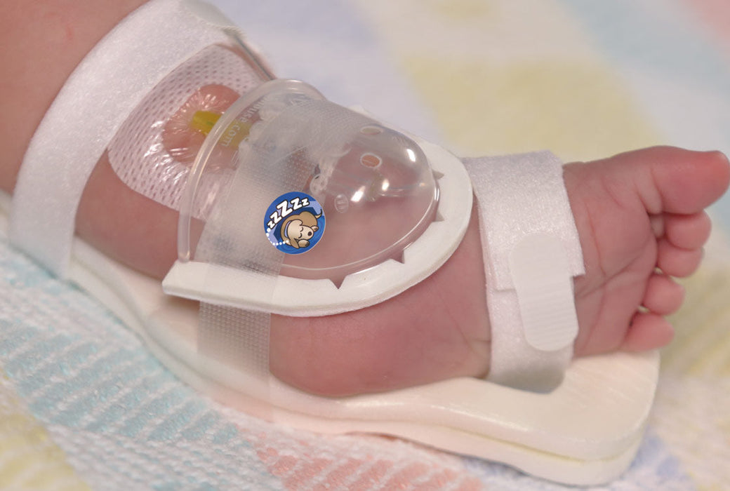 A baby's foot is secured in a white plastic device with straps. A small sticker shows a sleeping cartoon animal. The context is a hospital crib.