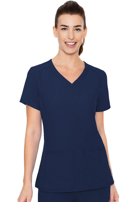 A woman in a navy blue v-neck scrub top stands against a white background. She is smiling.