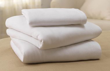 A neatly folded stack of white bed sheets rests on a light beige surface, possibly a bed or table, near a blurry off-white couch.