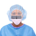 A woman, wearing a surgical cap, mask, and face shield, is looking directly at the viewer. She is dressed in light blue scrubs.
