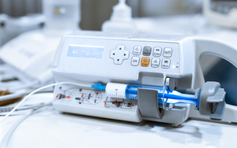 A medical syringe pump sits on a surface, displaying a digital readout and control panel. Buttons are labeled in Cyrillic script: Очист, Запуск, Болюс, О/б, ОК, Стоп, Очистка, Меню. The context is a clinical setting.