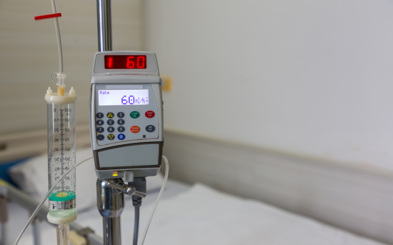 A medical infusion pump, displaying "60 l/h", is dispensing fluid from a graduated cylinder, attached to a pole in a hospital room.