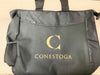 A black tote bag sits on a surface. The bag features a gold "C" and the word "CONESTOGA" embroidered on the front.