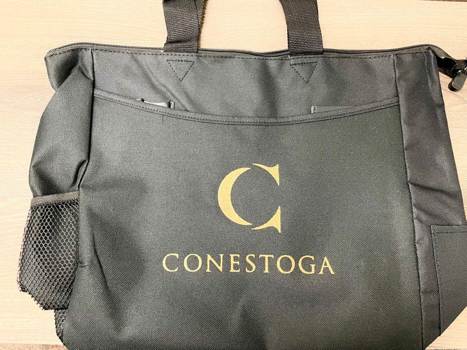 A black tote bag sits on a surface. The bag features a gold "C" and the word "CONESTOGA" embroidered on the front.