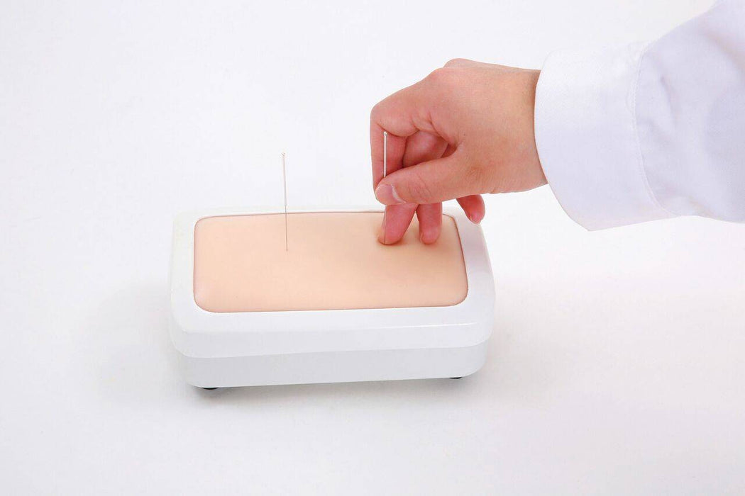 A hand inserts a needle into a skin-like surface on a rectangular training device. The device sits on a white surface.