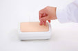 A hand inserts a needle into a skin-like surface on a rectangular training device. The device sits on a white surface.
