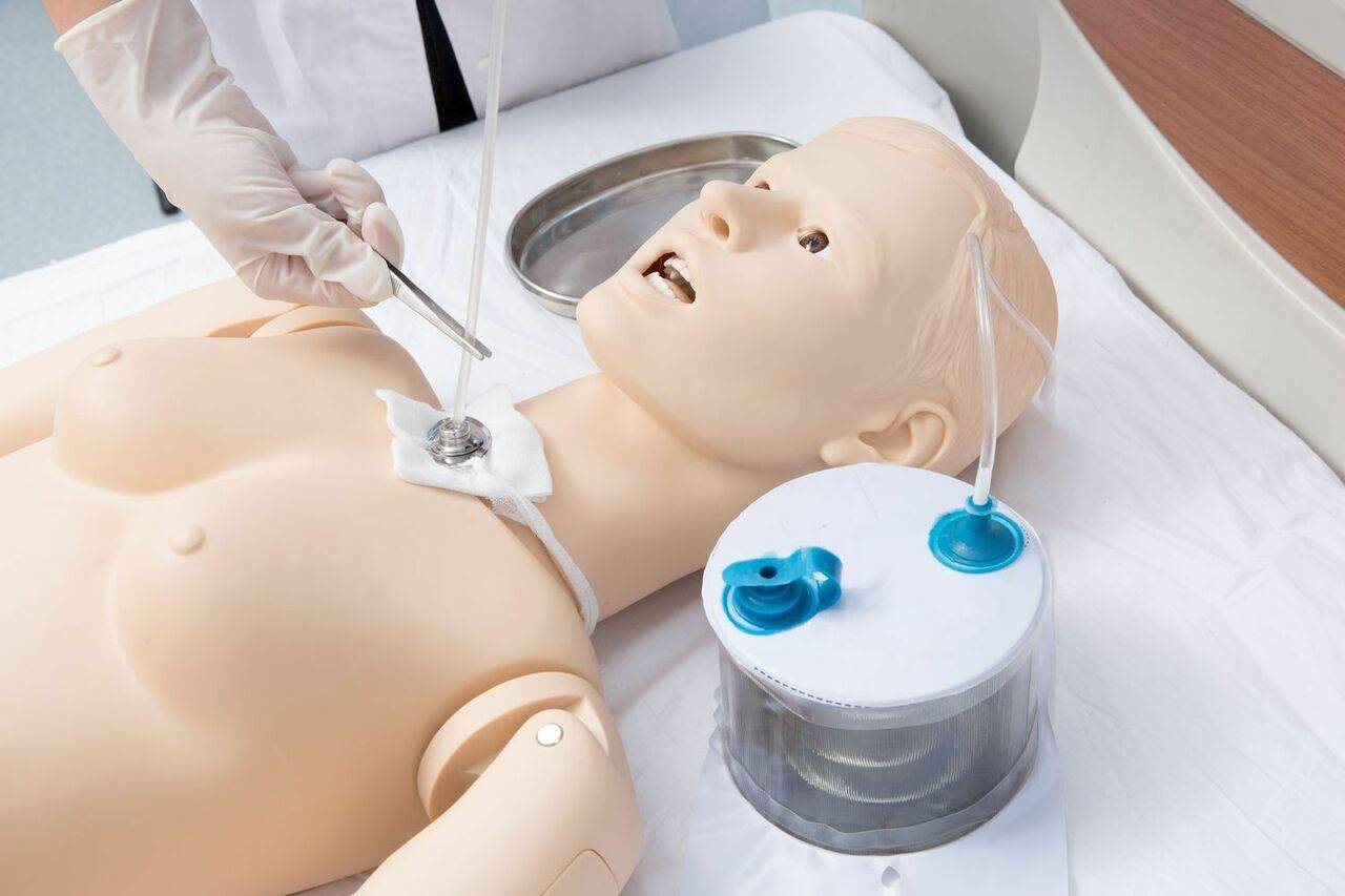 A medical mannequin lies supine; a gloved hand inserts a tube into its neck. The procedure takes place on a flat surface, near a small cylindrical container.